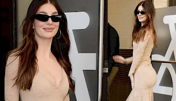 A woman with long brown hair wearing sunglasses and a fitted beige dress stands in front of a black and white background, smiling at the camera.