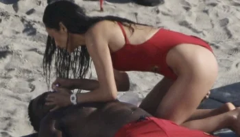 A woman in a red swimsuit kneels on the sand, giving first aid or CPR to a person lying on their back at the beach.