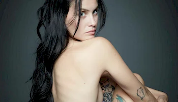 A woman with long dark hair sits turned slightly away, looking over her shoulder at the camera. She has visible tattoos on her arm and leg, and the background is a plain, dark color.