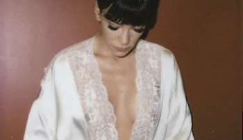 A woman with dark hair and bangs looks down while wearing a white satin robe with lace trim, against a plain brown background.