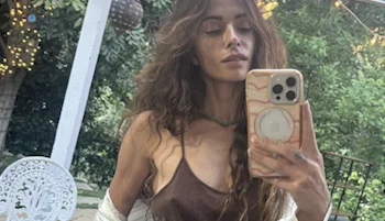 A woman with long wavy hair takes a selfie outdoors using a phone with a marble-patterned case. She wears a brown top and a light outer layer, with greenery and string lights in the background.