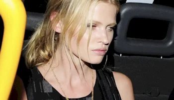 A woman with blonde hair, wearing a black sleeveless top and a gold necklace, sits in the backseat of a vehicle looking out the window with a serious expression.