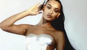 A woman with long dark hair poses against a plain background, wearing a shiny, strapless, metallic silver top and holding her hand to the side of her head.