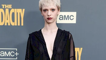 A person with short platinum blond hair wearing a black deep V-neck outfit poses in front of a grey backdrop with yellow “THE” and “AMC+” logos.