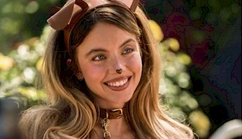A woman with long, wavy hair wears a brown dog ear headband, a brown collar with a gold tag, and has a heart-shaped nose painted on her face. She is smiling outdoors with greenery in the background.