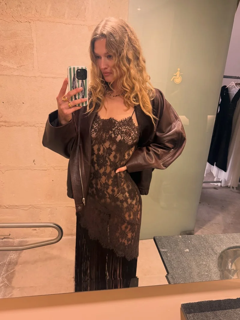 A woman with wavy blonde hair takes a mirror selfie, wearing a brown jacket over a lace black dress. She holds a phone with a striped case.