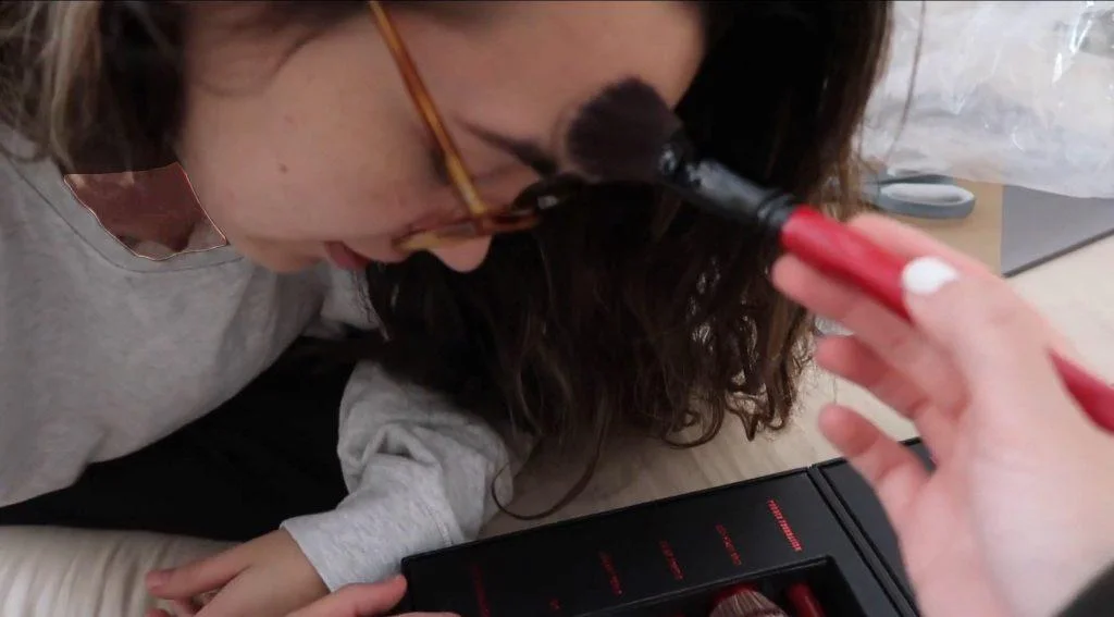 A girl wearing glasses leans forward as another hand applies makeup to their forehead with a brush. A makeup kit with red-handled brushes is visible in the foreground.