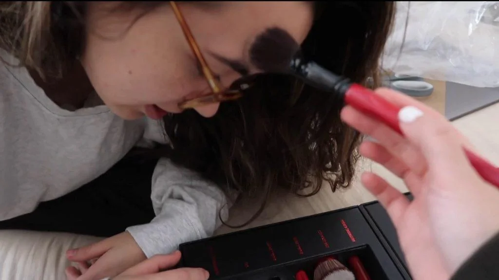 A girl wearing glasses leans forward as another hand applies makeup to their forehead with a brush. A makeup kit with red-handled brushes is visible in the foreground #2.