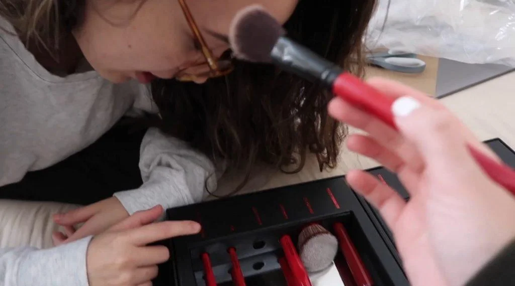 A girl wearing glasses leans forward as another hand applies makeup to their forehead with a brush. A makeup kit with red-handled brushes is visible in the foreground #3.