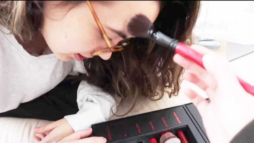 Brightened version of a girl wearing glasses leans forward as another hand applies makeup to their forehead with a brush. A makeup kit with red-handled brushes is visible in the foreground.