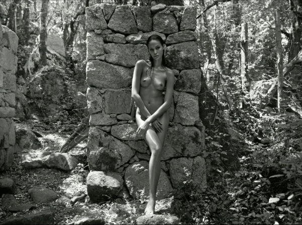 A naked woman leaning back against a ruined stone wall in a forest. The photo is black and white.