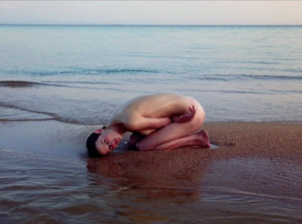 A nude woman curled up on wet sand at the shore, posing in a fetal position with their head resting on their arms, looking toward the camera. The calm sea and soft sky are visible in the background.