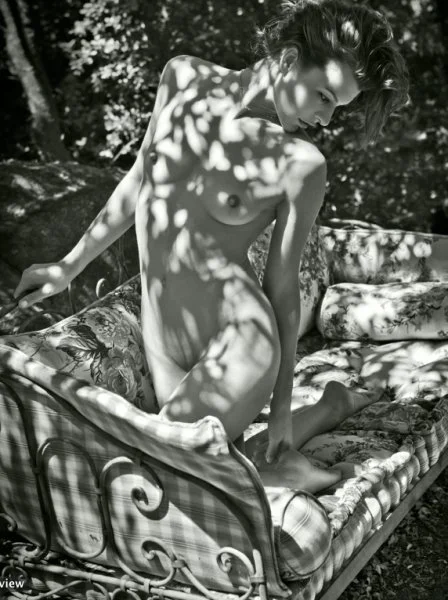 A naked woman kneel on a sofa that appears to be outdoors. Sun and shadows are on her body. The photo is black and white.