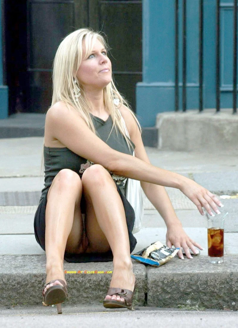 A woman with long blonde hair sits on a curb, wearing a dark dress and sandals, with a drink and a snack beside her. She looks upward and rests her arm on her knee.