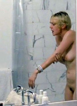 Side view of a naked and wet blonde girl with short hair standing inside a shower, holding up one of her arms using her other arm.