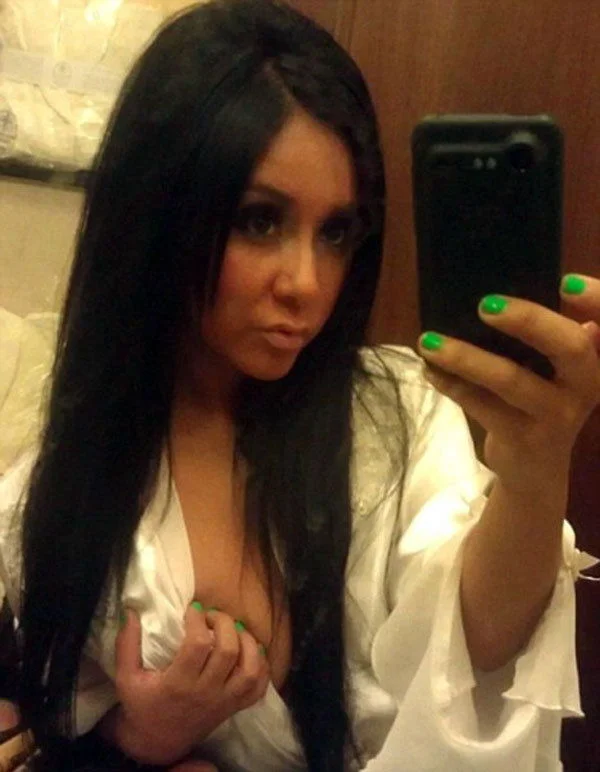 A woman with long dark hair wearing a white robe takes a selfie in a mirror. She has green nail polish and is slightly pouting at the camera. One side of her robe is pulled down, revealing part of her chest.