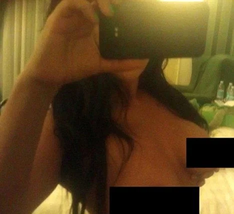 A woman takes a topless selfie, holding her phone in front of her face. Black bars cover her breasts.