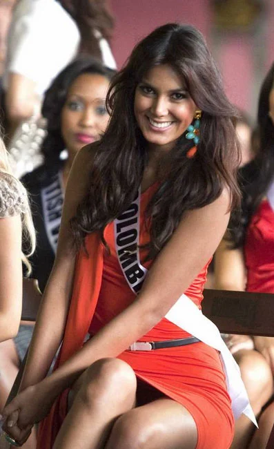A woman wearing a Colombia sash and a red dress sits smiling with her hands on her knees. Other women with pageant sashes sit nearby in the background.