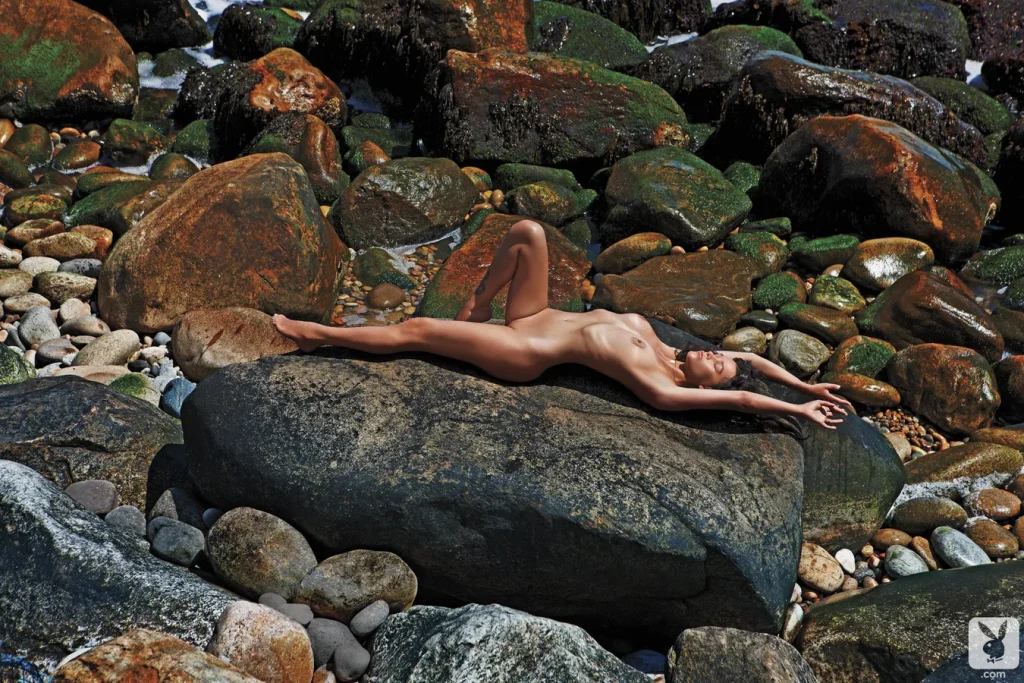 Far away side view of a naked woman lying on her back on a big rock at a rocky beach. The sun is shining.