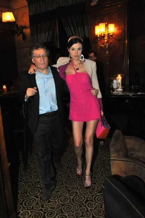 A man in a suit and glasses walks with a woman in a short pink dress and matching accessories inside a dimly lit, elegant room with patterned flooring and dark furnishings.