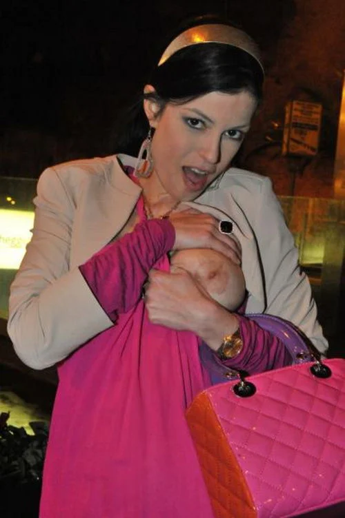 A woman wearing a pink jacket and large heart-shaped earrings touches one of her exposed breasts with both hands. She has mouth open and looks into the camera.
