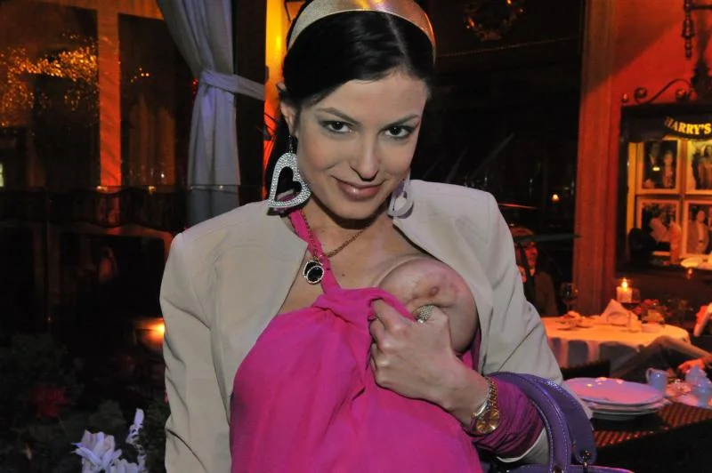 A woman wearing a pink jacket and large heart-shaped earrings squeezes one of her breasts that are half-way out of the pink dress she wears in a dimly lit restaurant. She smiles at the camera.