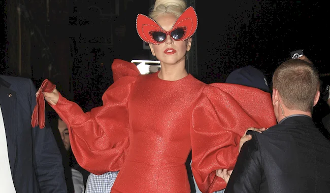 Lady Gaga wearing a bold red dress with large puffed sleeves, red gloves, and oversized red sunglasses shaped like butterfly wings, surrounded by people in a dark setting.