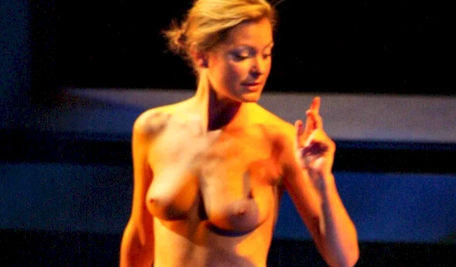 A naked blonde woman standing on a stage with lights and shadows on her body. She raises one arm as if to point.