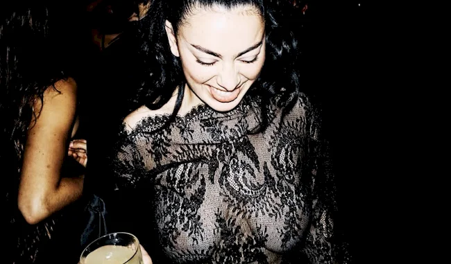 A woman in a sheer black lace dress smiles and looks down, holding a drink in one hand against a dark background.
