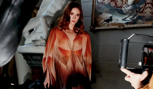 A woman with wavy auburn hair wears a flowing, pleated dress in earthy tones while standing indoors near a white statue and ornate painting; camera equipment and spotlights are visible around her.