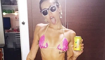 Close up of Miley Cyrus in a pink bikini poses indoors, sticking out her tongue and holding a yellow soda can. She wears sunglasses.