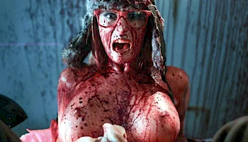 Close up of a topless woman wearing glasses and a fir hat. She is covered with blood and has her mouth open.
