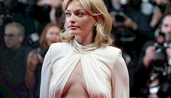 A woman with blonde hair wearing a white dress with a high neckline and cutout front stands on a red carpet, with photographers and people in the background.