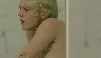 A woman with short blonde hair appears distressed while taking a shower, seen through a glass shower door with water droplets visible on the surface.