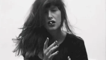 Black and white photo of a woman with long hair, eyes closed, slightly open mouth, and hands raised near their face, holding a cigarette in her mouth against a plain background.