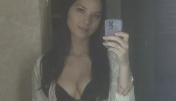 A woman with long dark hair is taking a mirror selfie with a phone. She is wearing a light-colored outer garment over a black top. The background is dimly lit.