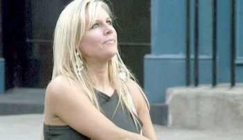 A woman with long blonde hair, wearing a sleeveless dark top and dangling earrings, looks upward while sitting outdoors near a concrete step and blue building.