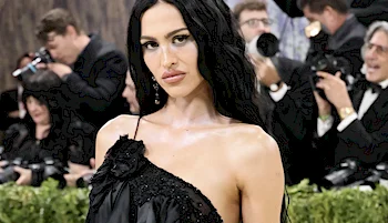 A woman with long black hair wears a one-shoulder black dress, posing confidently in front of photographers at a formal event.