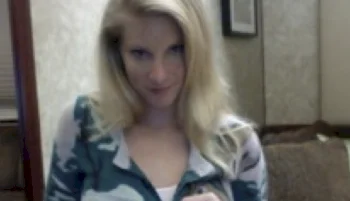 A woman with long blonde hair wearing a camo-patterned jacket stands indoors, looking directly at the camera with a slight smile. The background features beige walls and a brown couch.