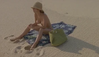 Far away view of a naked woman, wearing a hat and sitting on a towel at the beach.
