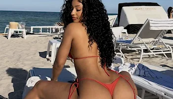 Black woman with long curly hair wearing a red bikini sits on a lounge chair at a sandy beach, with the ocean and more lounge chairs visible in the background.