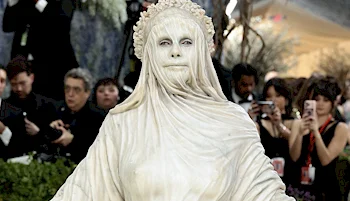A woman dressed as a marble statue, with white body paint, a draped toga-like costume, and a laurel crown, stands among photographers at a formal event.