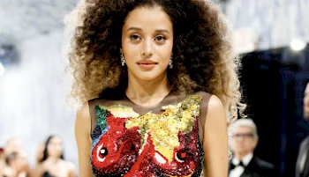 A woman with voluminous curly hair wears a sleeveless dress featuring a colorful, sequined fish design. She stands indoors at a formal event with blurred attendees in the background.
