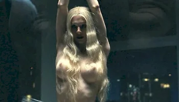 A topless blonde woman standing with arms raised. Light and shadows are on her body.