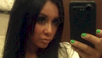 A woman with long dark hair takes a selfie in a mirror, holding a black phone with one hand showing bright green nail polish. The background appears to be a bathroom.