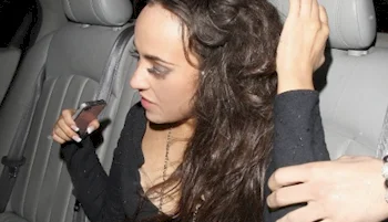 A woman with long brown hair sits in the back seat of a car, holding a phone in one hand and brushing back her hair with the other. She is wearing a black top and appears to be looking downward.
