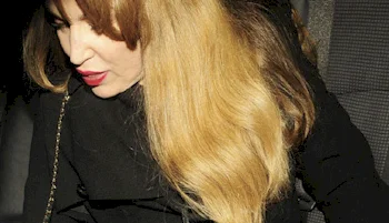 A woman with long, light brown hair and red lipstick looks downward while sitting in a car, wearing a black coat.