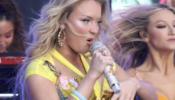 A woman with long blonde hair in a yellow outfit sings into a sparkly microphone on stage. She wears large earrings and colorful bracelets, with backup dancers visible behind her.