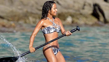 A woman in a patterned bikini, smiling while paddleboarding in clear blue water near rocky shorelines.