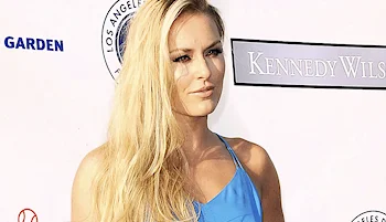 A woman with long blonde hair wearing a blue dress stands in front of a white backdrop with logos and text, including KENNEDY WILSON and LOS ANGELES.